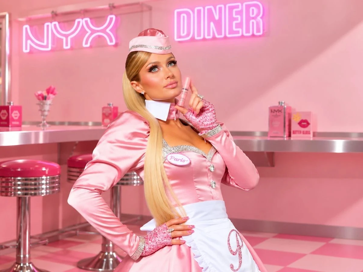 Paris Hilton en tenue de serveuse rose dans un diner aux néons NYX, un Butter Gloss à la main