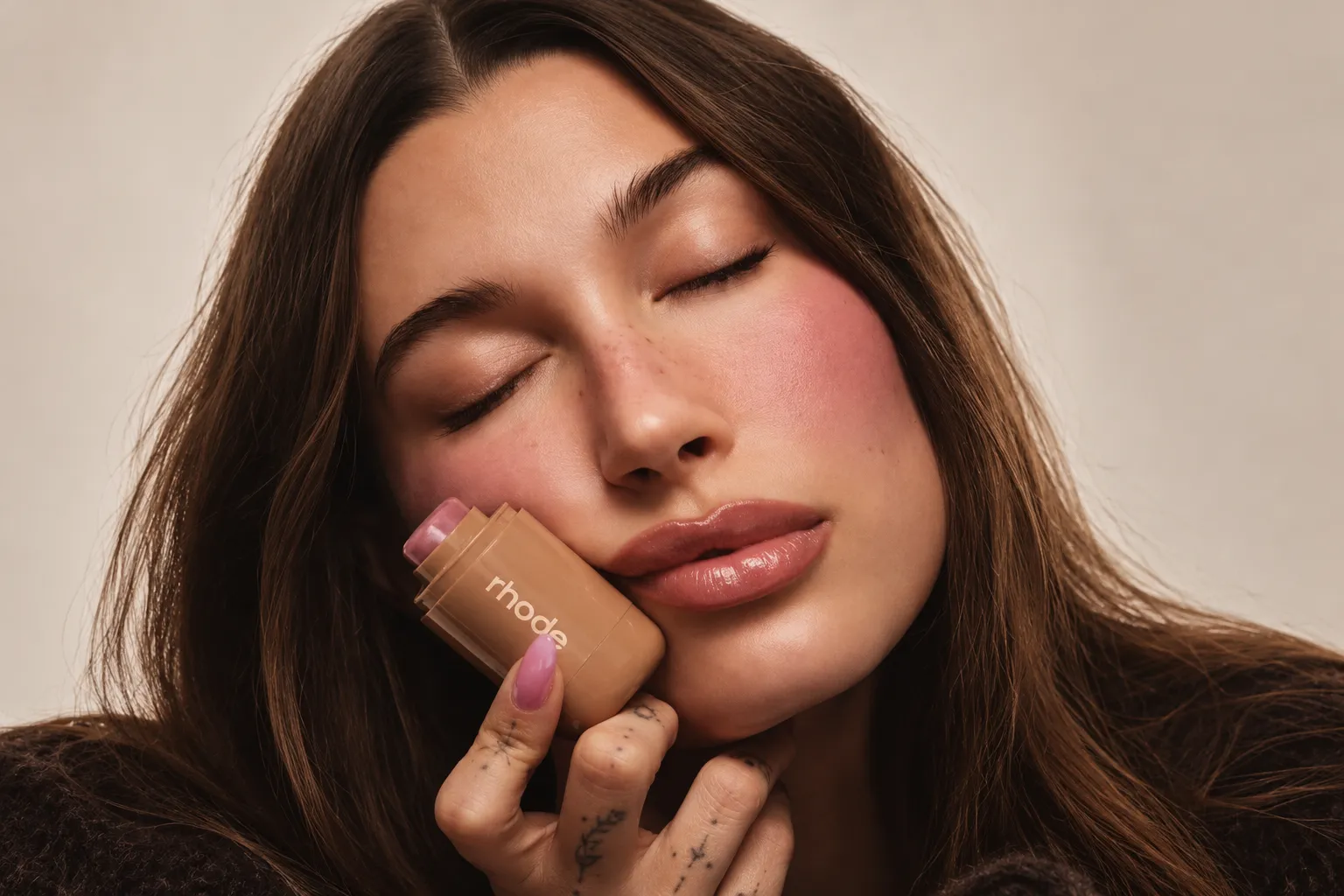 Jeune femme aux yeux fermés tenant un stick de blush Rhode contre sa joue